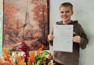 Na zdjęciu uczeń naszej szkoły z listem gratulacyjnym. Obok na sztaludze stoi obraz, poniżej jesienne kwiaty w wazonie.