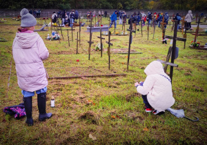 Na cmentarzu widać krzyże drewniane wbite w ziemie. Na pierwszym planie widać dwie dziewczynki, jedna po lewej stronie stoi, druga po prawej kuca.