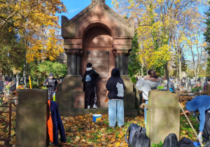 Na cmentarzu przy grobie stoją uczniowie. Porządkują miejsce.