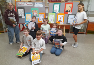 Na szkolnym korytarzu na tle tablicy z portretami Tuwima stoją dzieci nagrodzone w szkolnym konkursie.