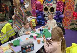Trzy dziewczynki siedzą przy stole. Na stole rozłożone są przybory papiernicze. W tle widać maskę meksykańską z kolorowymi balonami. Po lewej stronie kadru widać osobę prowadzącą zajęcia.