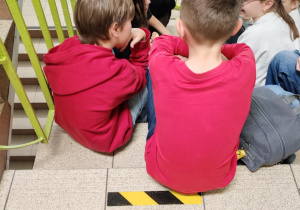 Na schodach siedzi dwójka dzieci ubrana na czerwono. Po lewej stronie kadru widać poręcz.