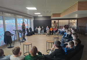 W pomieszczeniu dzieci siedzą w półkolu. Po lewej stronie kadru widać osobę prowadzącą zajęcia. Za oknami widać trybuny stadionu Widzewa.