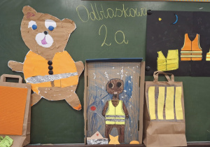 Na zielonej tablicy od lewej wisi miś w kamizelce odblaskowej, kolejny miś oraz kamizelki odblaskowe wykonane z papieru kolorowego.