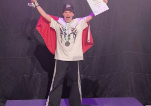 Na drugim stopniu podiumm stoi nasz uczeń. Na ramionach ma flagę Polski, ręce uniesione do góry. W lewej ręce trzyma dyplom, w prawej statuetkę.