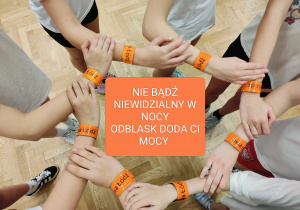 Na zdjęciu w okręgu stoją uczennica. Mają splecione ręce. Na nadgarstkach widać opaski odblaskowe. W środku zdjęcia znajduje się pomarańczowy kwadrat z napisem "nie bądź niewidzialny w nocy odblask doda ci mocy"