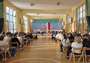 Na sali gimnastycznej siedzą uczniowie ubrani na galowo. W głębi kadru w oddali widać grupę dzieci prowadzącą apel.