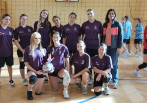 Na sali gimnastycznej na tle siatki stoi drużyna dziewcząt wraz z trenerką