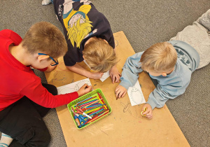 Na podłodze przy kartonie widać trzech chłopców. Dwóch leży na brzuchu na podłodze, jeden siedzi. Rysują plakat.