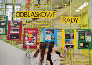 Na kracie na szkolnym korytarzu wiszą prace uczniów wykonane na lekcjach informatyki. Nad pracami wisi napis "Odblaskowe rady". Na środku kadru siedzą dwie dziewczynki i rękami wskazują na plakaty.