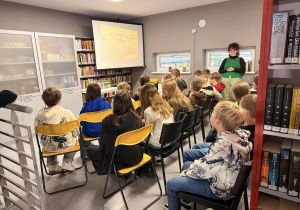 W małym pomieszczeniu biblioteki uczniowie siedzą na krzesełkach. Widać osobę prowadzącą zajęcia oraz regały z książkami.