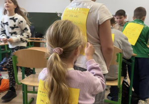 Dwie dziewczynki mają przyklejone do pleców żółte kartki, na których są napisane miłe słowa. Jedna z dziewczynek pisze drugiej miłe słowa.