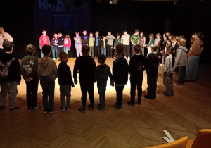 W pomieszczeniu uczniowie stoją w dużym okręgu. Trzymają się za ręce.