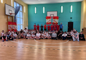 Na sali gimnastycznej zdjęcie grupowe uczestników spotkania z drużyną Widzew Futsal. Uczniowie ubrani są w stroje sportowe. Na sali rozwieszone są emblematy klubu sportowego Widzew. Widać również nauczycieli wf.