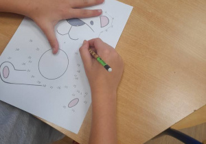 Na stole leży kartka z narysowanym misiem. Widać ręce dziecka rysującego misia metodą "połącz kropki"