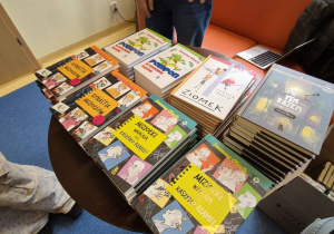 Na stole leżą książki. Widać trzy osoby stojące wokół stołu. Z tyłu widoczna jest pomarańczowa kanapa.