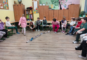 W sali lekcyjnej dzieci siedzą na krzesłach w kręgu. Na podłodze stoją ułożone buty jeden za drugim.