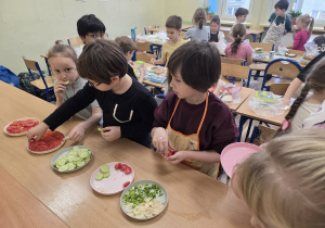 Dzieci siedzą przy ławkach. Na blatach stoją talerze z kolorowymi kanapkami