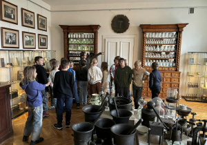 W sali muzeum stoją dzieci, oglądają eksponaty, słuchają osoby prowadzącej zajęcia.