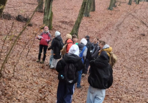 Na zdjęciu grupa dzieci idących lasem. Widać drzewa i podłoże przykryte liśćmi.