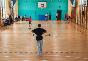 Na sali gimnastycznej widać chłopców stojących na parkiecie. Są rozstawieni do gry w piłkę nożną. Widać akcję toczącą się w prawym górnym rogu zdjęcia.