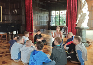 Na podłodze w sali muzealnej w kręgu siedzą uczniowie. Widać osobę prowadzącą zajęcia.