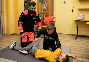 W sali lekcyjnej na podłodze na macie leży chłopiec. Drugi chłopiec pod kierunkiem ratownika medycznego układa kolegę w pozycji bocznej.