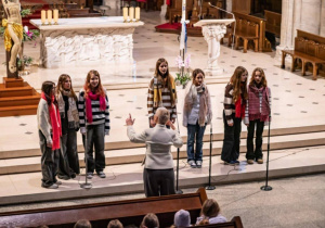 W kościele na schodach w prezbiterium stoją uczennice naszej szkoły. Przed nimi stoi nauczycielka nasze szkoły i dyryguje śpiewem.