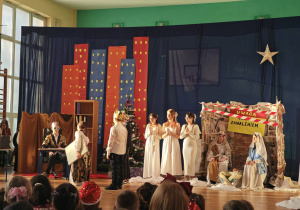 Na zdjęciu uczniowie grający w jasełkach. Widać trzech królów, Heroda, aniołki, Marię z Józefem i dzieciątkiem. W tle dekoracja, na której widać wieżowce oraz stary budynek.