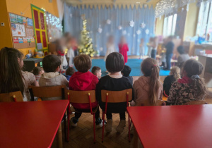 W sali siedzą uczniowie z naszej szkoły. Oglądają przedstawienie. W tle widać dekorację oraz dzieci biorące w przedstawieniu. Twarze dzieci są wyszarzone ze względu na ochronę danych osobowych.