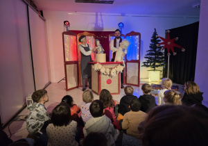 Uczniowie siedzą na podłodze, oglądają przedstwienie. Widać dwie osoby trzymające w rękach lalki. W tle świąteczna dekoracja.