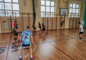 Na sali gimnastycznej uczniowie stoją w różnych miejscach. Trwa rozgrywka w piłkę nożną.