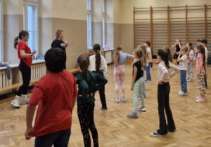 Na sali gimnastycznej dzieci stoją w różnych miejscach. Patrzą na osoby prowadzące zajęcia.