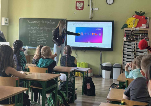 W sali szkolnej uczniowie siedzą w ławkach. Widać osobę prowadzącą zajęcia. Korzysta ona z tablicy multimedialnej.