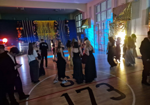 Na sali gimnastycznej uczniowie ubrani w stroje balowe stoją na parkiecie.