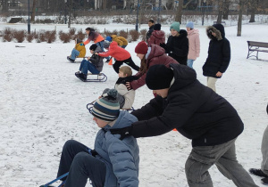 W parku na śniegu widać uczniów siedzących na sankach oraz uczniów stojących za nimi i trzymających ich za ramię.