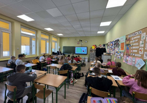 Uczniowie siedzą w sali lekcyjnej. W głębi widać osobę prowadzącą zajęcia siedzącą przy biurku.