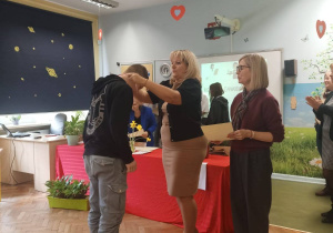 W sali lekcyjnej przed stołem przykrytym czerwonym materiałem stoją organizatorzy konkursu. Wręczają medal naszemu uczniowi.