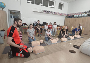W sali szkolnej pod ścianą na krzesełkach siedzą uczniowie. Na podłodze rozłożone są fantomy. Po lewej stronie kadru widać osobę prowadzącą zajęcia. Przy fantomach klęczą uczniowie.