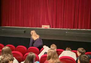 Na widowni siedzą uczniowie. W głębi kadru widać kawałek sceny zasłoniętej czerwoną kurtyną.