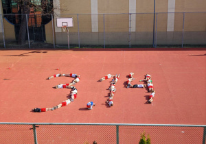 Widok boiska szkolnego z góry. Na boisku leżą uczniowie. Są ułożeni w kształcie liczby 3,14