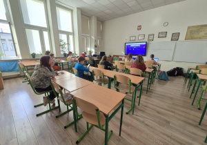 W sali lekcyjnej uczniowie siedzą w ławkach. Na ekranie telewizora wyświetlone jest zadanie konkursowe.