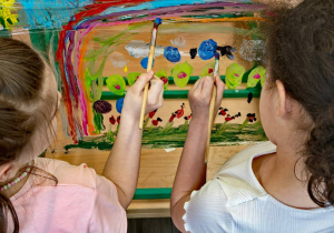 Dwie dziewczynki rysują wiosenny obrazek na folii spożywczej zawiniętej na nogi krzesła.