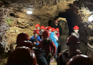 W korytarzu kopalni stoją uczniowie. Na głowach mają czerwone kaski.