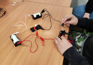 Na ławce leżą żarówki, wiatraczki, brzęczyki połączone w obwody elektryczne. Widać też ręce dziecka.