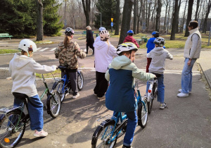 Grupa dzieci siedzi na rowerach. Na głowach mają kaski. Po prawej stronie kadru widać osobę prowadzącą zajęcia, które odbywają się na alejkach na motodromie.