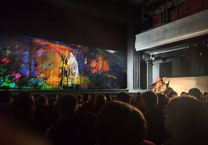 Na widowni w teatrze widać siedzące dzieci. W głębi widać wyświetloną grafikę sceniczną. W głębi po prawej stronie widać aktora grającego w przedstawieniu trzymającego topór.