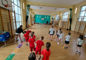 W sali gimnastycznej widać dwie drużyny dzieci ustawionych rzędem. Drużyna po lewej stronie jest ubrana w czerwone stroje, drużyna po prawej stronie w białe koszulki i granatowe krótkie spodenki.