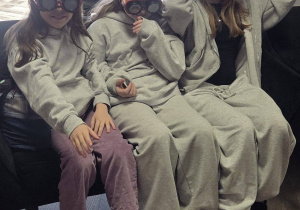 W pomieszczeniu na fotelach siedzą trzy dziewczynki. Na głowę mają założone specjalne okulary.