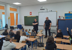 Uczniowie siedzą w ławkach w klasie. W głębi widać przedstawicieli Straży Miejskiej prowadzących zajęcia.